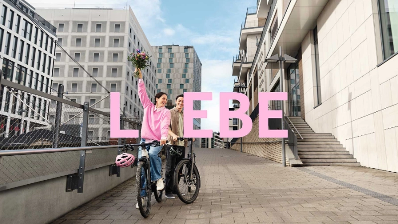 2 JobRadler:innen auf einem Fahrrad in einem urbanen Umfeld mit einem Blumenstrauß.